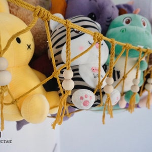 May include: A yellow, white, and black striped stuffed animal, a green stuffed dinosaur, and a yellow stuffed bunny are hanging in a macrame hammock with wooden beads. The hammock is made of yellow rope and has tassels.