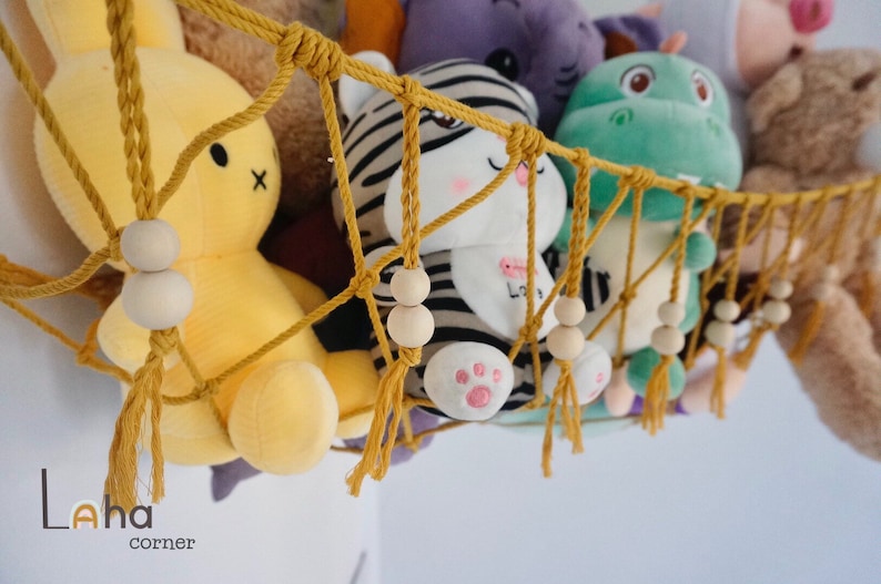 May include: A yellow, white, and black striped stuffed animal, a green stuffed dinosaur, and a yellow stuffed bunny are hanging in a macrame hammock with wooden beads. The hammock is made of yellow rope and has tassels.