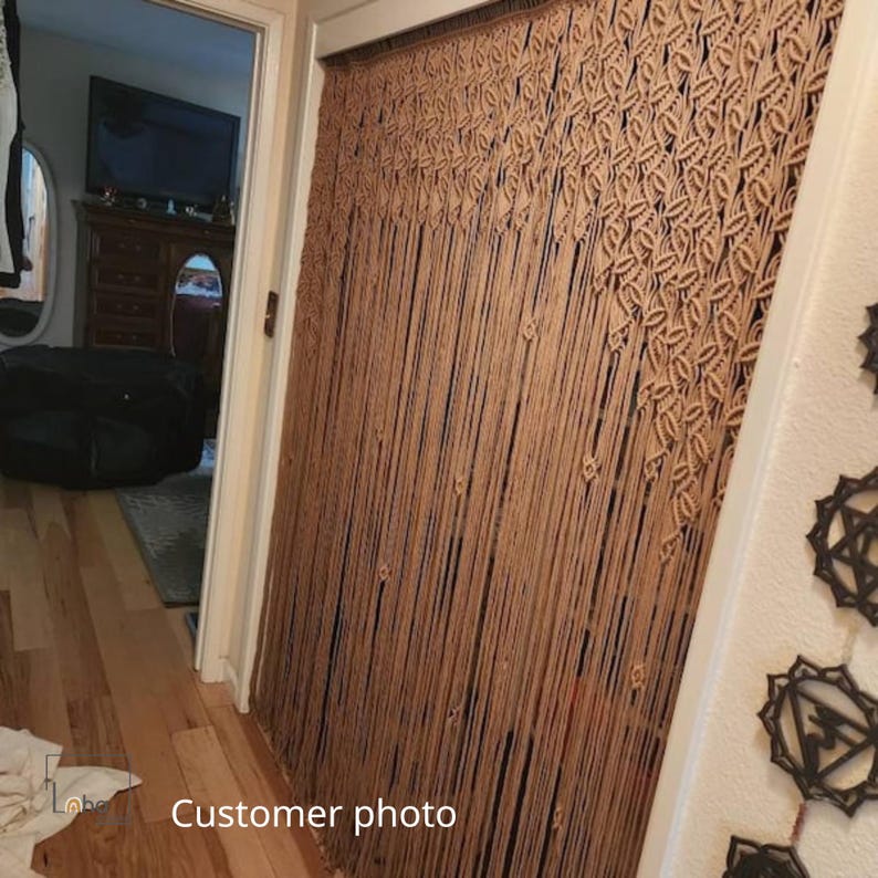 May include: A beige macrame door curtain featuring an intricate knotted pattern at the top that transitions into long, vertical hanging cords. The curtain covers a doorway, displaying a dense, textured design made from woven cotton rope.