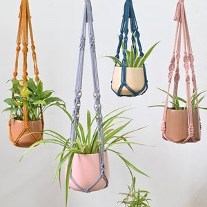 May include: Four macrame plant hangers with different colored cords and wooden beads. The hangers are holding potted plants. The hangers are hanging from a wooden rod.