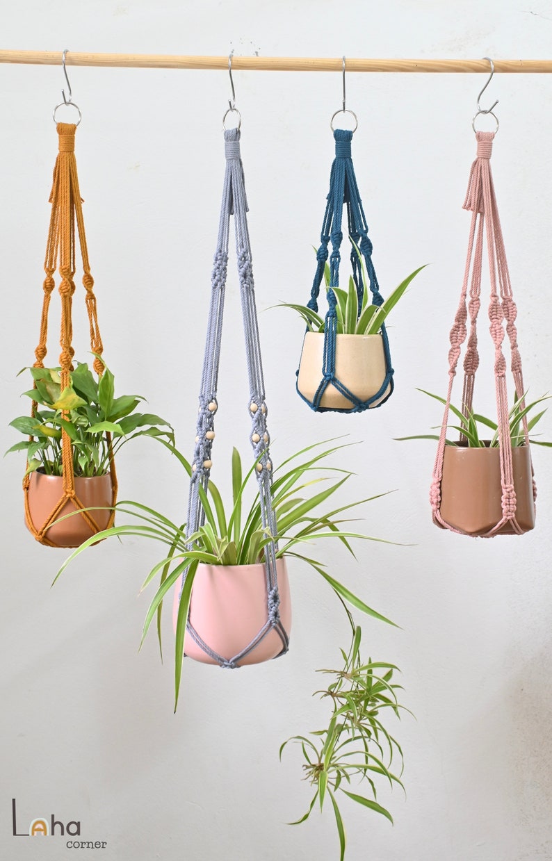 May include: Four macrame plant hangers with different colored cords and wooden beads. The hangers are holding potted plants. The hangers are hanging from a wooden rod.