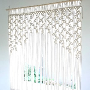 May include: An off-white macrame wall hanging or curtain suspended from a wooden dowel. The design features intricate knotted leaf patterns arranged in cascading diagonal sections, transitioning into long, straight vertical fringe cords.