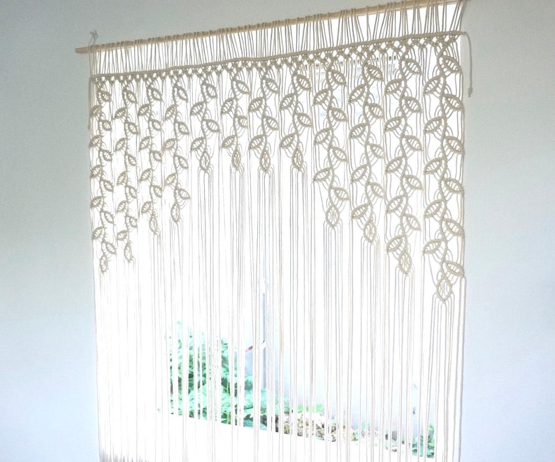 May include: An off-white macrame wall hanging or curtain suspended from a wooden dowel. The design features intricate knotted leaf patterns arranged in cascading diagonal sections, transitioning into long, straight vertical fringe cords.