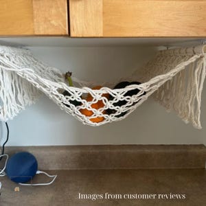 Puede incluir: Una hamaca de frutas de macram&eacute; color crema, suspendida debajo de un armario, contiene naranjas, aguacates y otros productos. El dise&ntilde;o tejido presenta una red anudada con bordes con flecos. El texto "Images from customer reviews" est&aacute; en la parte inferior.