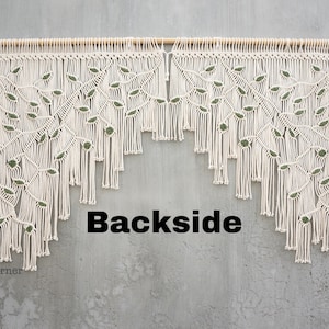 May include: A macrame wall hanging featuring off-white cotton cord knotted into a branch and leaf pattern with small dark green accents. The piece is suspended from a horizontal wooden dowel and includes the text Backside and Laha corner.