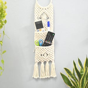 Op de afbeelding: Een crèmekleurige macramé wandorganizer met een ronde ophanglus, twee gestapelde vakken met een ruitpatroon en vier kwastjes aan de onderkant. De vakken bevatten een rekenmachine, een blauwe stift, een schaar en een afstandsbediening.