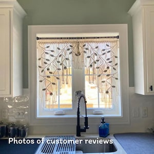 May include: A cream-colored macrame window valance featuring an intricate leaf-like pattern with knotted fringe details. The textile hanging is suspended from a dark rod and displays a symmetrical design of woven cord loops and vertical strands.