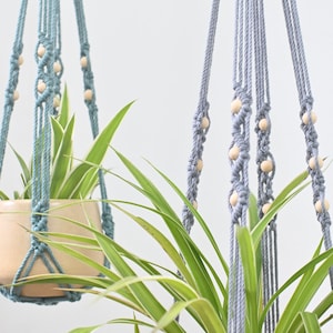 May include: Two macrame plant hangers with wooden beads, one in light blue and one in gray, hanging from the ceiling with a plant in each.