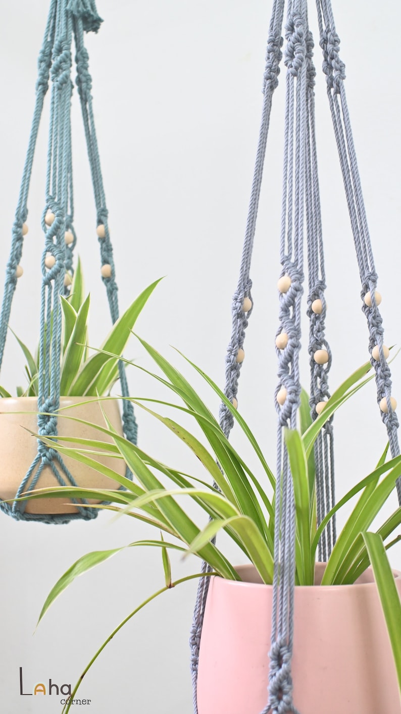 May include: Two macrame plant hangers with wooden beads, one in light blue and one in gray, hanging from the ceiling with a plant in each.