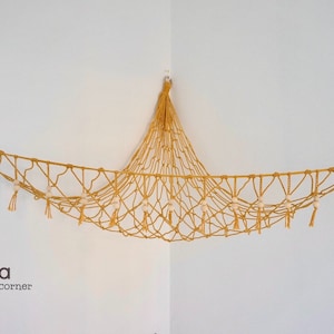 May include: A yellow macrame hammock with white beads and tassels hanging from the ceiling in a corner. The hammock is decorated with two rainbow wall hangings.