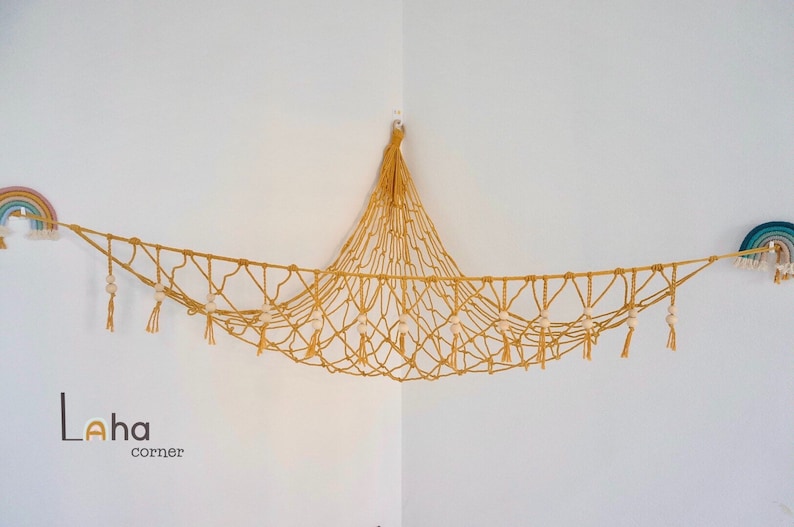 May include: A yellow macrame hammock with white beads and tassels hanging from the ceiling in a corner. The hammock is decorated with two rainbow wall hangings.