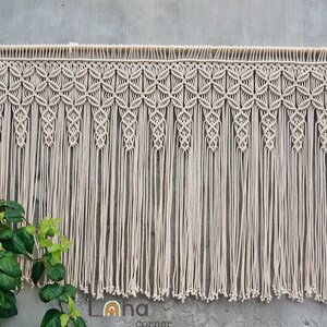 May include: A cream-colored macrame wall hanging suspended from a horizontal wooden dowel. The piece features an intricate top section with repeating geometric knot patterns, transitioning into long, vertical fringe strands with knotted ends.