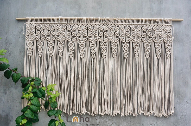 May include: A cream-colored macrame wall hanging suspended from a horizontal wooden dowel. The piece features an intricate top section with repeating geometric knot patterns, transitioning into long, vertical fringe strands with knotted ends.