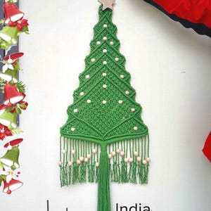 May include: A green macrame wall hanging shaped like a Christmas tree, featuring a wooden star at the top and light-colored wooden beads integrated into the knotted cord design. The piece includes a long central tassel and fringe at the base.
