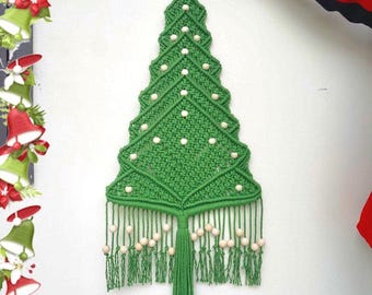 May include: A green macrame wall hanging shaped like a Christmas tree, featuring a wooden star at the top and light-colored wooden beads integrated into the knotted cord design. The piece includes a long central tassel and fringe at the base.