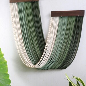 May include: A wall hanging featuring two dark brown wooden bars suspending long, draped strands of cord in shades of cream, sage green, and dark forest green, accented by a single string of small, round, light-coloured wooden beads.