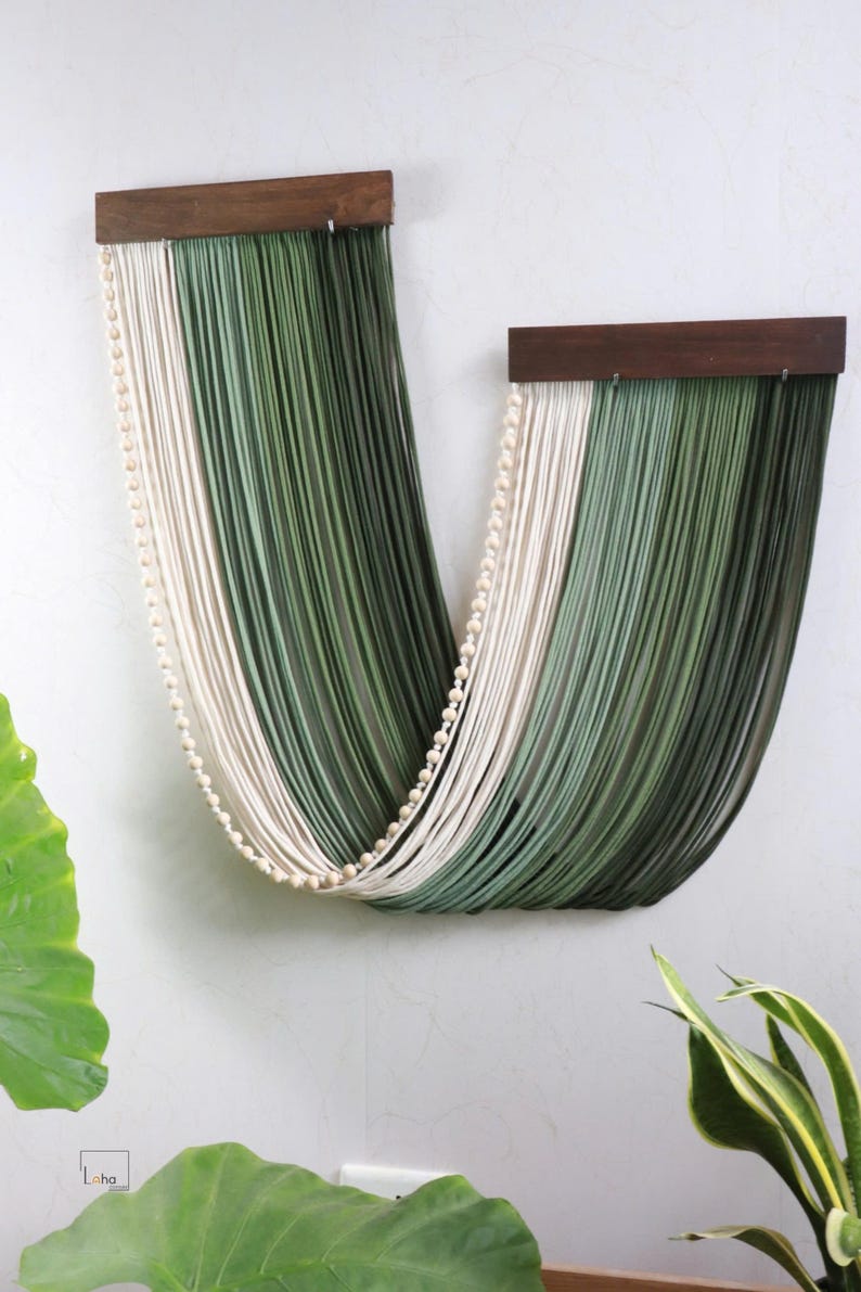 May include: A wall hanging featuring two dark brown wooden bars suspending long, draped strands of cord in shades of cream, sage green, and dark forest green, accented by a single string of small, round, light-coloured wooden beads.
