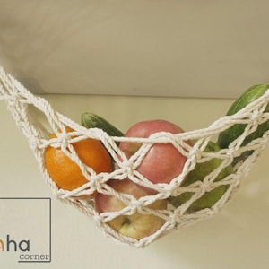Puede incluir: Una hamaca de frutas de macram&eacute; color crema, suspendida del techo, llena de frutas y verduras frescas. La hamaca contiene naranjas, manzanas y pepinos. El logotipo de Laha Corner es visible en la esquina inferior izquierda.