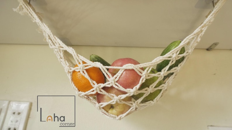 Puede incluir: Una hamaca de frutas de macram&eacute; color crema, suspendida del techo, llena de frutas y verduras frescas. La hamaca contiene naranjas, manzanas y pepinos. El logotipo de Laha Corner es visible en la esquina inferior izquierda.