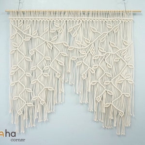 May include: A white macrame wall hanging with a leaf pattern. The hanging is made of cotton cord and has a wooden dowel at the top.