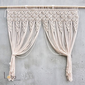 May include: An off-white macrame curtain hanging from a wooden rod, featuring a complex knotted geometric pattern at the top and long, flowing vertical fringe strands that are gathered into two distinct sections with knotted ends.