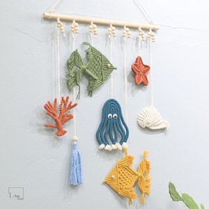 May include: A wall hanging with a nautical theme featuring a variety of crocheted sea creatures, including a starfish, an octopus, a seashell, and two fish. The hanging is made with colorful yarn and includes a wooden dowel with beads.