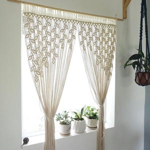 May include: A cream-colored macrame curtain featuring a repeating leaf-like knot pattern. The curtain hangs from a wooden rod and is gathered at the sides with matching cord ties, creating a decorative window covering with long, vertical fringe.