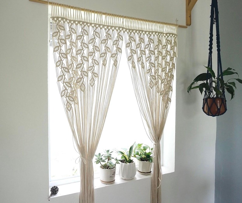 May include: A cream-colored macrame curtain featuring a repeating leaf-like knot pattern. The curtain hangs from a wooden rod and is gathered at the sides with matching cord ties, creating a decorative window covering with long, vertical fringe.