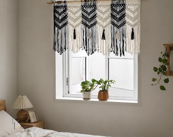 May include: A macrame window valance featuring alternating vertical panels of black and cream-colored cord. The design includes chevron patterns and long, dangling fringe with small tassels at the bottom, suspended from a wooden rod.