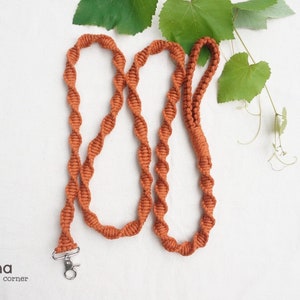 Puede incluir: Una correa de macram&eacute; color &oacute;xido con un patr&oacute;n de nudos en espiral continuo y un mosquet&oacute;n giratorio de metal plateado en un extremo. La correa incluye un asa en forma de bucle y tiene el texto Laha corner en la esquina inferior izquierda.