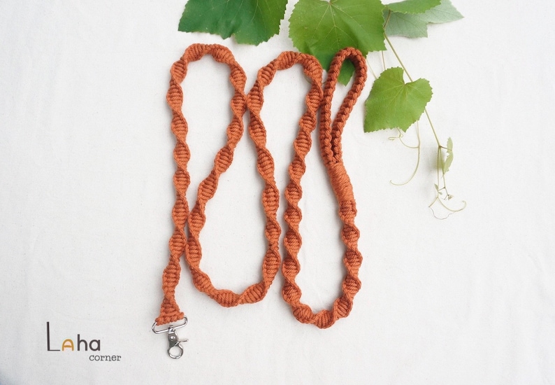 Puede incluir: Una correa de macram&eacute; color &oacute;xido con un patr&oacute;n de nudos en espiral continuo y un mosquet&oacute;n giratorio de metal plateado en un extremo. La correa incluye un asa en forma de bucle y tiene el texto Laha corner en la esquina inferior izquierda.