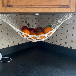 Puede incluir: Una hamaca de frutas de macram&eacute; blanco llena de naranjas, tomates y otros productos. La hamaca est&aacute; suspendida debajo de un mueble de madera, contra un salpicadero de azulejos. La encimera de la cocina es negra.