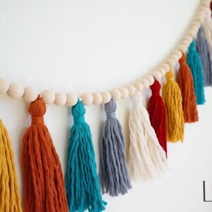 May include: A colorful tassel garland with wooden beads. The garland features tassels in shades of red, yellow, orange, teal, gray, and white.