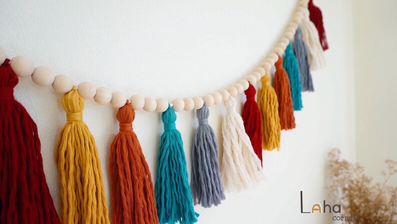 May include: A colorful tassel garland with wooden beads. The garland features tassels in shades of red, yellow, orange, teal, gray, and white.