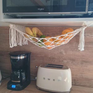 Cute white rope fruit hammock neatly hanging under a kitchen cabinet, holding bananas, oranges, and limes to keep the kitchen tidy and stylish.