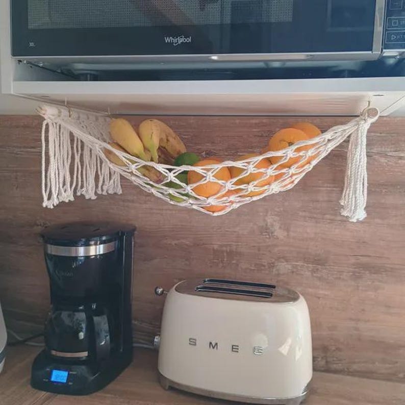 Cute white rope fruit hammock neatly hanging under a kitchen cabinet, holding bananas, oranges, and limes to keep the kitchen tidy and stylish.