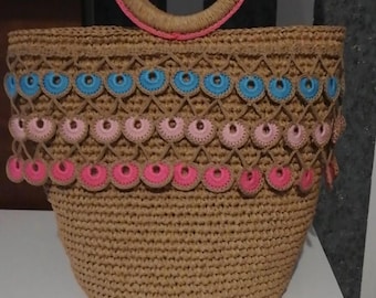 Puede incluir: Un bolso de mano de crochet marrón con un ribete de crochet rosa y azul. El bolso tiene un asa redonda y tejida.