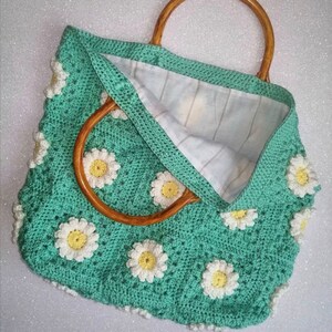 May include: A green crocheted tote bag with a wooden handle. The bag is decorated with white daisy flowers.