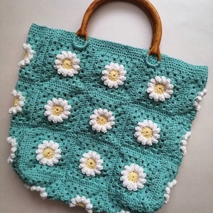 May include: A teal crocheted tote bag with a wooden handle and white daisy flower accents.