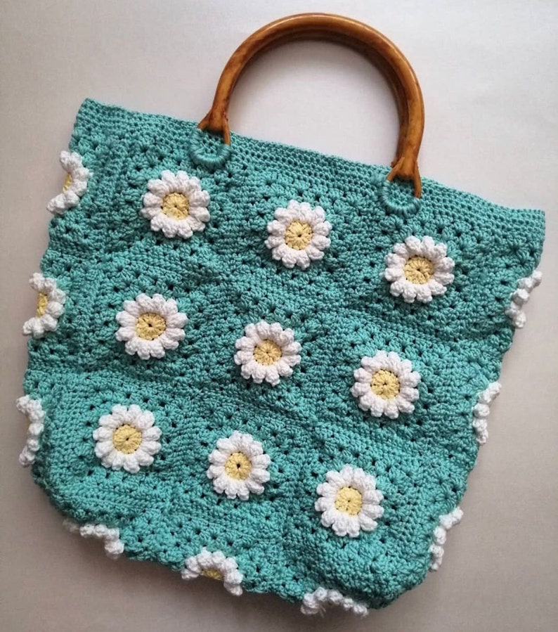 May include: A teal crocheted tote bag with a wooden handle and white daisy flower accents.