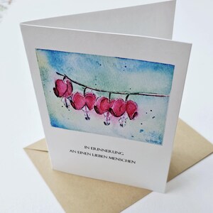 Puede incluir: Tarjeta blanca plegada con una pintura de acuarela de flores de corazón sangrante rosas sobre un fondo azul y verde. La tarjeta incluye el texto "IN ERINNERUNG AN EINEN LIEBEN MENSCHEN" en negro.