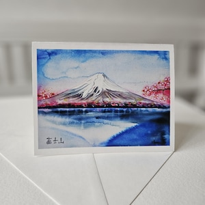 May include: Watercolor painting of Mount Fuji with a reflection in a lake. The mountain is snow-capped and the lake is blue. There are pink cherry blossoms in the foreground. The text "富士山" is written in Japanese at the bottom of the card.