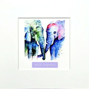 Könnte beinhalten: Gerahmtes Aquarellgemälde mit zwei Elefanten, deren Rüssel sich berühren. Die Elefanten sind in Blau-, Grün- und Rosatönen gemalt. Der Text "Stick Together" befindet sich unterhalb des Bildes in einem lila Rechteck.