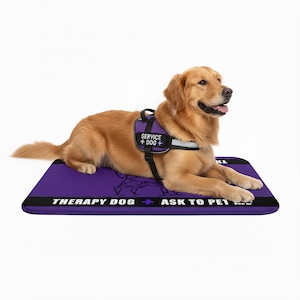 Op de afbeelding: Een golden retriever ligt op een rechthoekige paarse mat met zwarte randen. De mat bevat de witte tekst "THERAPY DOG + ASK TO PET ME". De hond draagt een paars tuigje met een patch met de witte tekst "SERVICE + DOG +" erop.