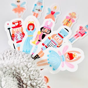 May include: A collection of colorful stickers featuring characters from The Nutcracker ballet. The stickers include ballerinas in pink and blue tutus, and toy soldiers in red and blue uniforms. The stickers are on a white background.
