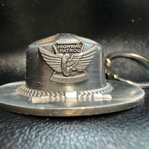 May include: Silver metal keychain shaped like a hat with the words "Highway Patrol" and a winged emblem on the front.