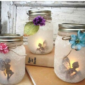May include: Three glass jars with fairy silhouettes inside, decorated with glitter, twine, and flowers. The jars are lit from within, creating a warm glow.