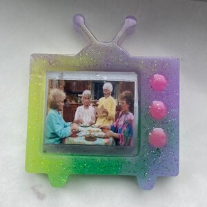 May include: A small, glittery, rainbow-colored television with a picture of four women sitting at a table. The television has three pink buttons on the side.