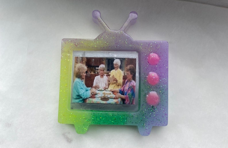 May include: A small, glittery, rainbow-colored television with a picture of four women sitting at a table. The television has three pink buttons on the side.