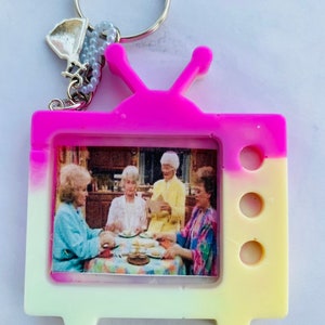 May include: A pink and yellow television-shaped keychain with a picture of four women inside. The keychain has a silver charm attached to it.